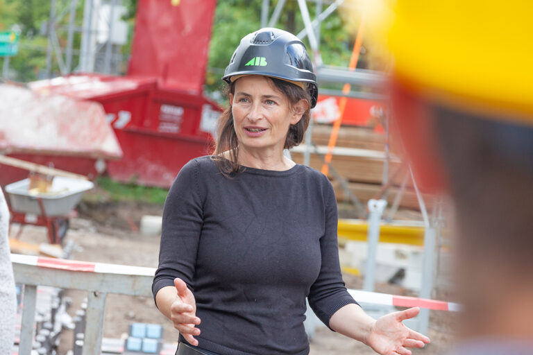 Dieses Bild dient der Darstellung der Carbonbetontage, damit sich Interessierte einen Eindruck machen können. Es zeigt eine Nahaufnahme einer Frau auf einer Baustelle. Sie trägt einen Helm und ein schwarzes Langarm-Shirt.